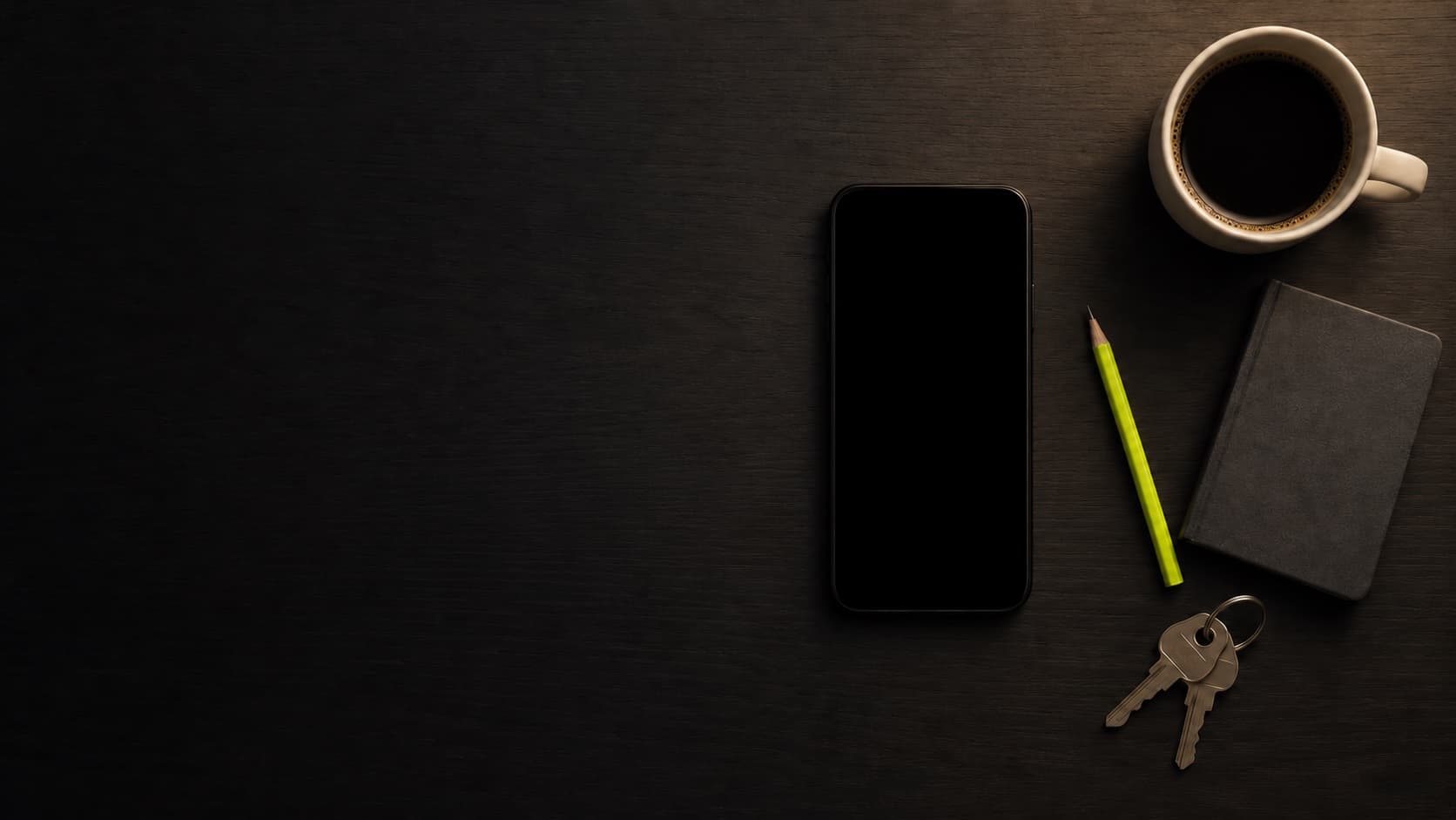 A phone lying flat on a dark desk, beside a coffee cup, a notebook, a pencil and keys.
